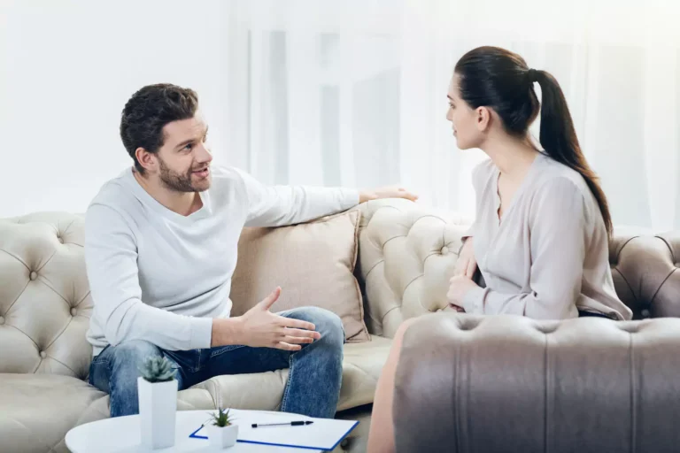 A husband and wife sitting on a couch talking to each other. There are four types of communication to strengthen your marriage.