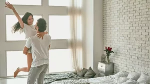 A man spinning his wife in the air by their bed. She is smiling because he knows how to please her sexually after consulting the Bible.