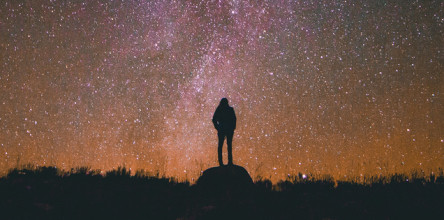 Silhouette of person standing on a hill looking into a beautiful night sky that's full of stars