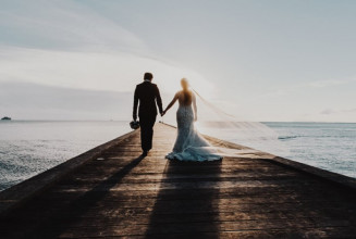 Shown from behind, a newlywed couple walking down a long pier over a lake into the sunset
