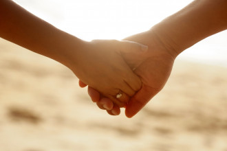 Close-up of husband holding wife's hand in front of an ocean sunset