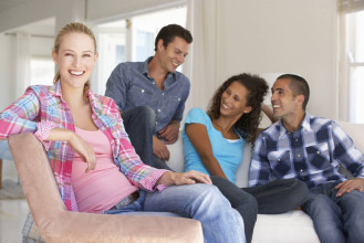 Two smiling couples hanging out in a living room, with one of the women in the foreground looking into the camera