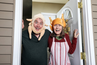 Couple overexuberant about Thanksgiving; wearing turkey hats as they stand in the front door welcoming visiting family
