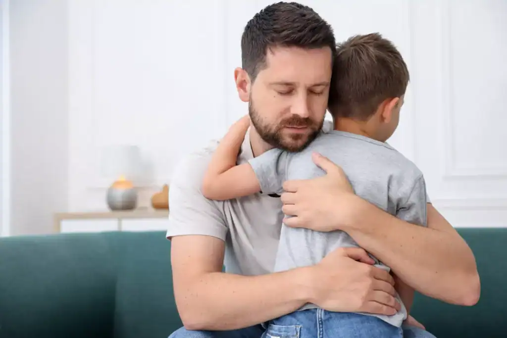 An adult and a child sit on a couch indoors, with the child being held in a close embrace. The image is often used to represent themes of single-parenthood and caregiving.