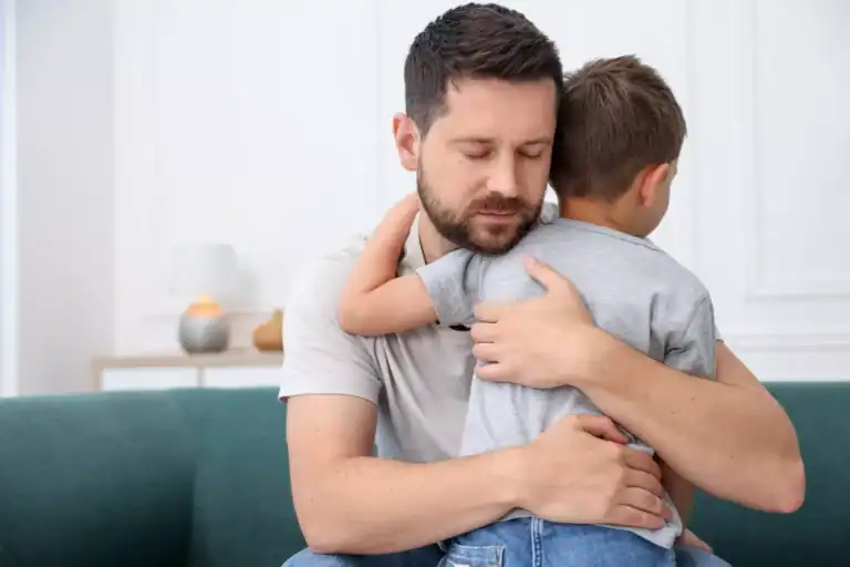 An adult and a child sit on a couch indoors, with the child being held in a close embrace. The image is often used to represent themes of single-parenthood and caregiving.