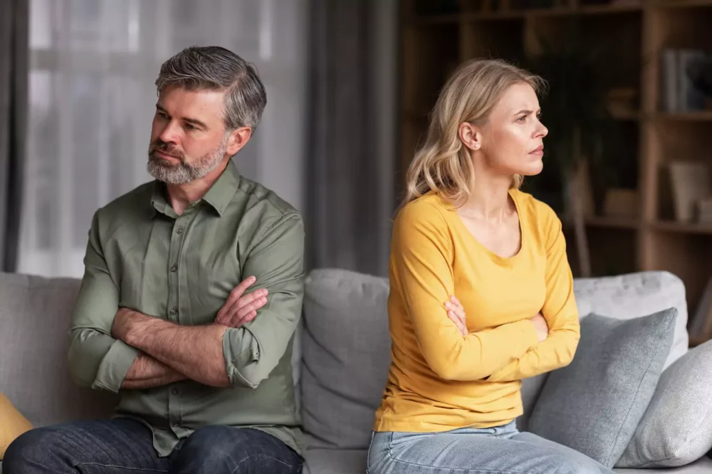 A middle aged couple sits on the couch with back turned to each other and arms crossed. Learning to deal with marriage conflict is important.