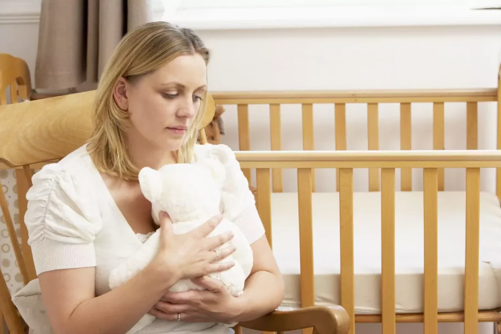 A grieving woman sitting in a nursery, holding a white teddy bear, symbolizing the emotional journey of coping with miscarriage.