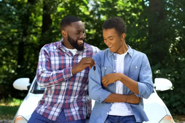 father and son sitting on the hood of a car. The dad is giving his son the keys to the car