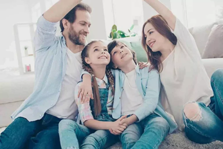 A happy family of four sits together on the floor of their bright and cozy living room. The parents, a smiling couple, raise their arms playfully, forming an arch over their two children, who gaze up at them with joy. The image reflects love, unity, and the lasting impact of strong relationships, symbolizing the importance of your legacy as a married couple