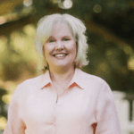 Karen Crum stands smiling against a background of trees.