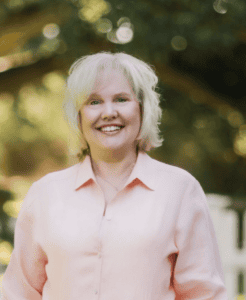 Karen Crum stands smiling against a background of trees.