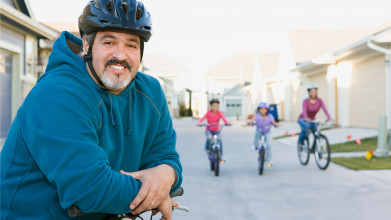 Mom, dad, and two kids out for a bike ride. Dad is stopped in the foreground, posing and smiling for the camera.