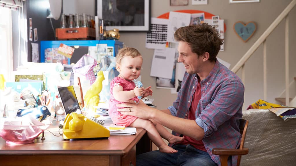 Young, smiling father balancing his toddler daughter who’s sitting on the edge of his desk