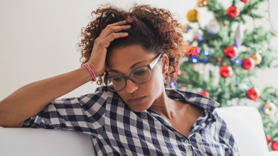 Unhappy-looking African-American woman sitting on her couch, resting her head in her hand; a Christmas tree in the background