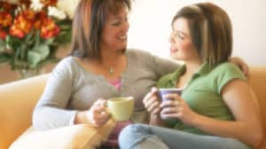 Mother and daughter sitting on sofa, looking at each other; mother has her arm around her daughter