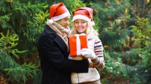 Couple going on a Christmas date, both wearing Santa hats, and the man passing the woman a red wrapped present