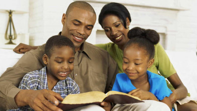 Smiling African-American family with mom, dad, and two young kids sitting on their couch, reading a Bible