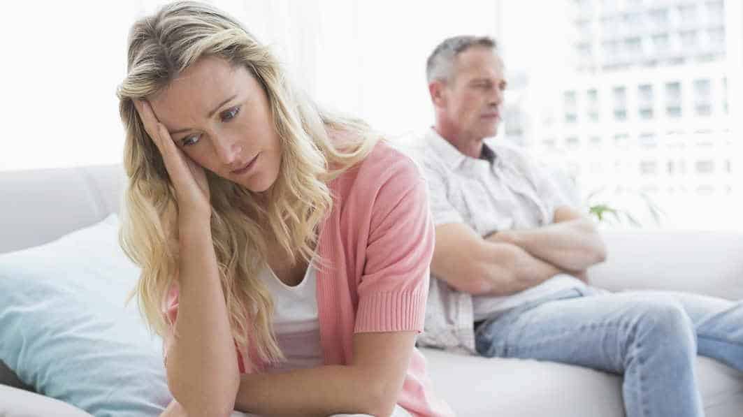 Wife looking down after a tense conversation with her husband, who is sitting arms crossed in the background in the background