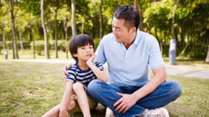 Man and his son sitting in the grass in a park. The father has his arm around his son.