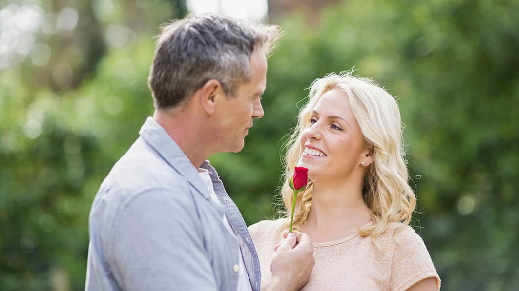 Middle-age couple standing together outside. He’s affectionately holding a rose up to her face.