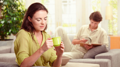 Woman in green holding a coffee cup, with her husband out of focus behind her reading a book