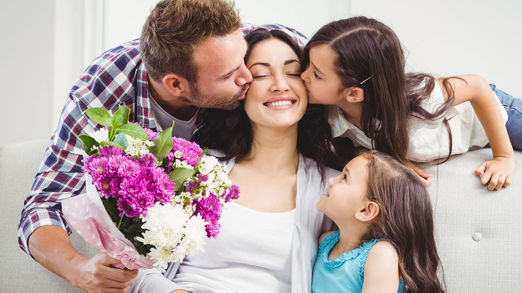 A mother surrounded by her family: two daughters and her husband kissing her on the cheek and holding a flower bouquet
