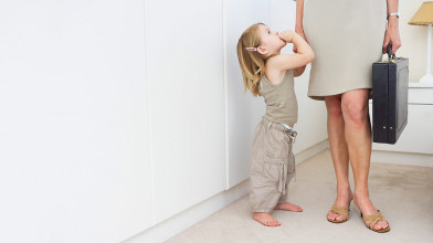 Young, pensive girl looking up at her mom who’s holding a briefcase and dressed professionally, as if she’s leaving for work