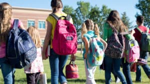 Shown from behind, a group of school children with backpacks walking across a green grass field