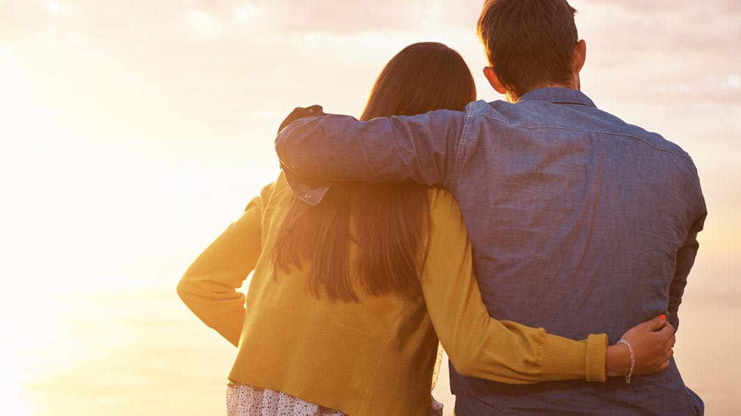 Couple sitting together with their arms around each other, gazing out at beautiful sunset