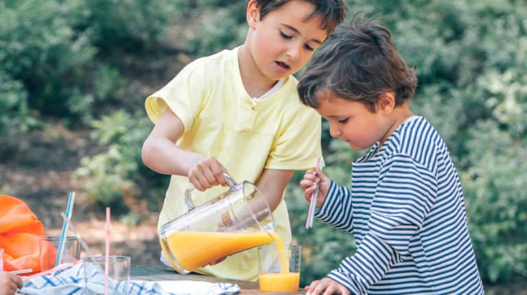 A young boy pouring a glass of orange juice for another boy at an outside table