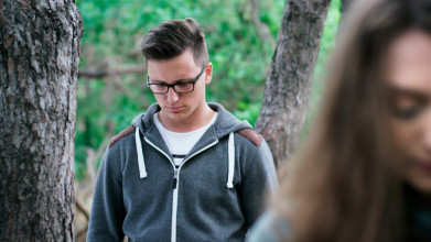 Troubled young couple standing apart outside, with focus on him in the background and her face blurry in the foreground
