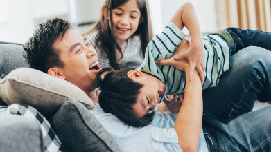 Laughing Asian father playfully wrestling with his young son and daughter on the couch