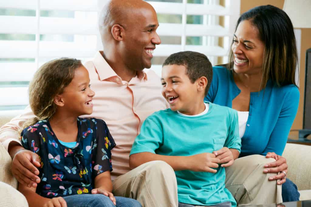 Dad, mom, and young son and daughter all laughing as they sit on the couch together