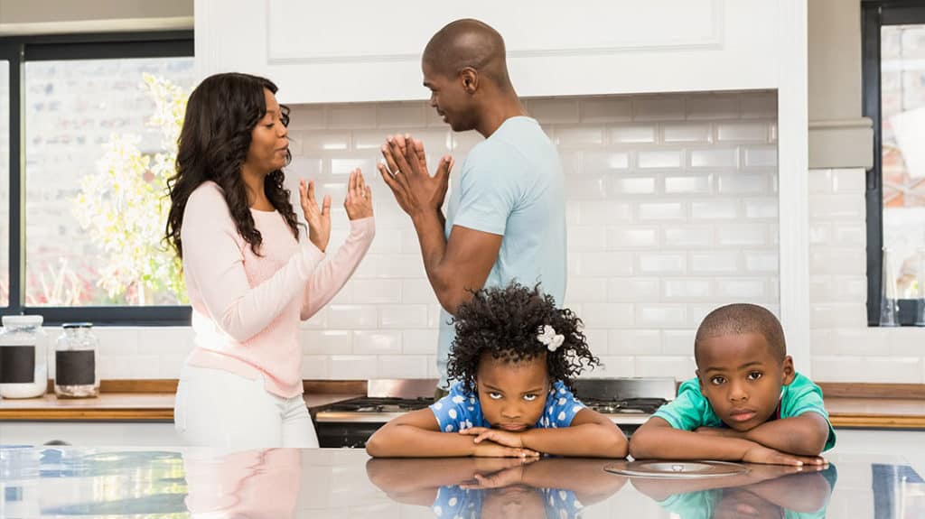 Young, unhappy brother and sister resting their heads on kitchen table while parents have serious discussion the background