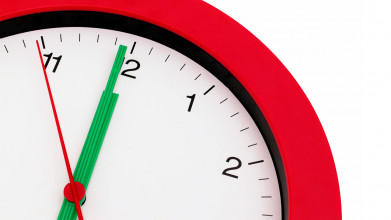Close up of a circular red clock with a white face, showing the time of about 5 seconds before noon (or midnight)