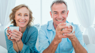 Happy middle-age couple sitting together on their couch, holding cups of coffee and smiling for the camera