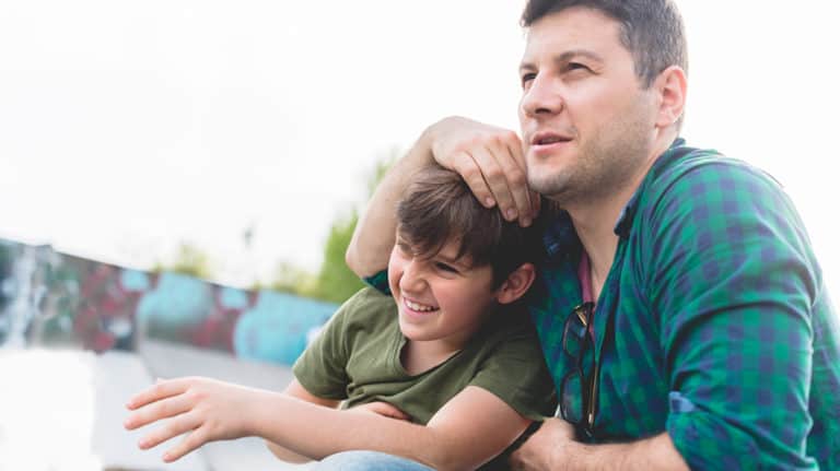 Dad has his arm affectionately wrapped around his smiling, young son’s head as they sit together outside