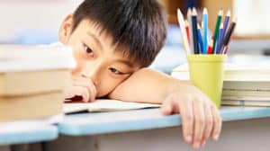 Young, serious-looking Asian boy resting his head on his arm on his school desk