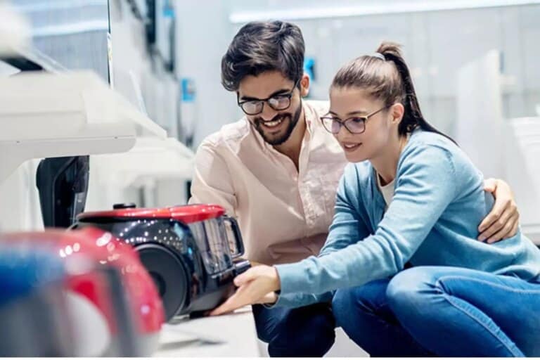 A smiling couple is shopping for home appliances together in a well-lit electronics store. They are closely examining a red and black vacuum cleaner, with the man wrapping his arm around the woman in a supportive gesture. Their shared focus and engagement suggest the importance of financial alignment and decision-making as a couple, highlighting how to get on the same page financially.