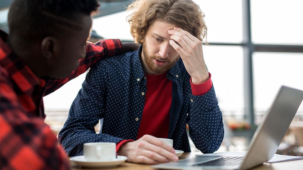 Two men sitting in a coffee shop. One looks troubled, and the other has his arm on his shoulder to comfort him.