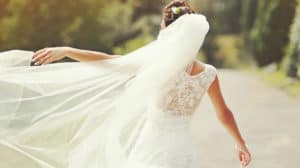 Shown from behind, a bride standing outside, her left arm raised, following the flow of her veil in the breeze