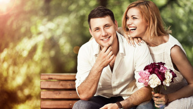 Happy couple sitting on a park bench. He’s smiling at the camera, she’s laughing, looking to the side and holding a bouquet