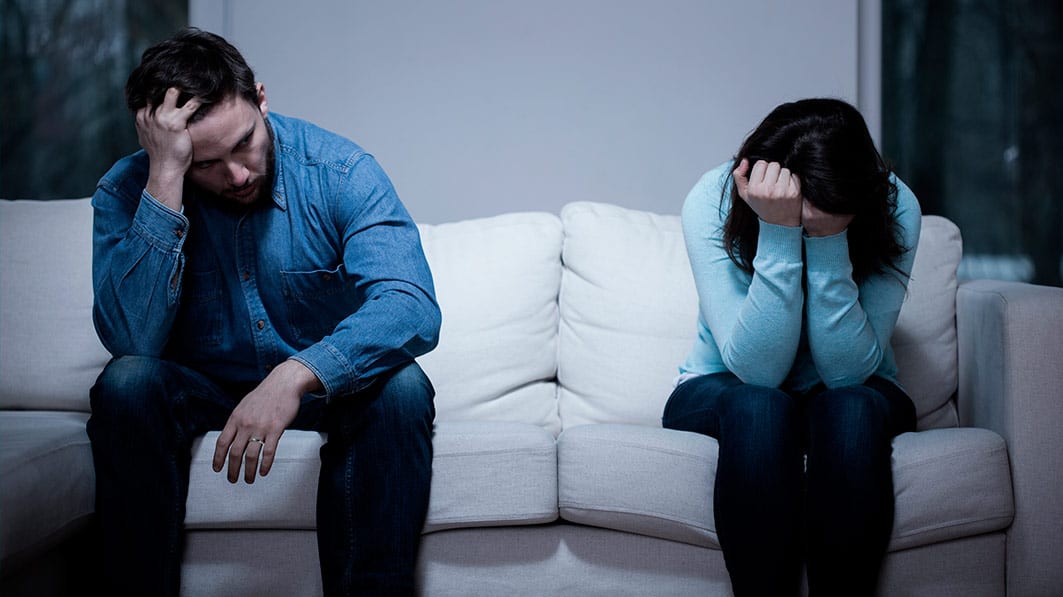 An upset husband and wife sitting apart on a couch with their heads in their hands