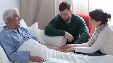 Adult children sitting beside their elderly father who is lying in bed