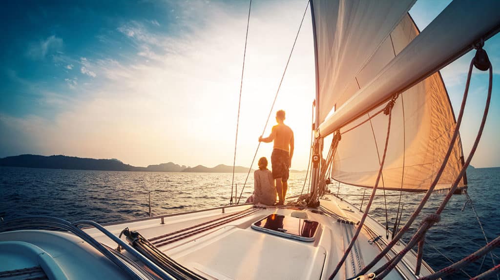 Shown from behind, a couple on the bow of a sailboat looking out over the ocean at a sunrise