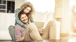 Happy couple; husband sitting, holding a tablet with his feet propped up, wife standing, hugging him from behind