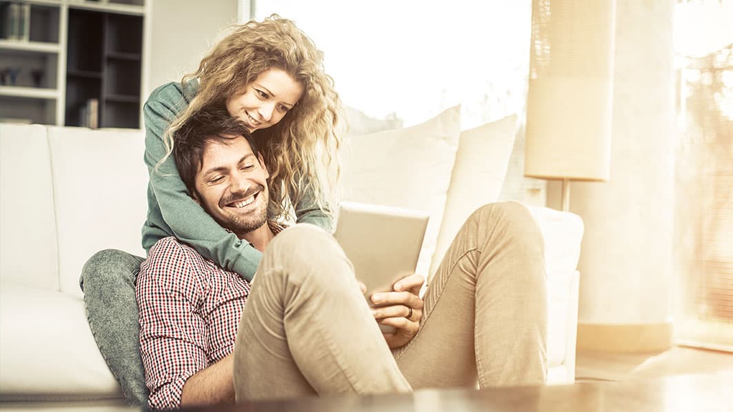 Happy couple; husband sitting, holding a tablet with his feet propped up, wife standing, hugging him from behind