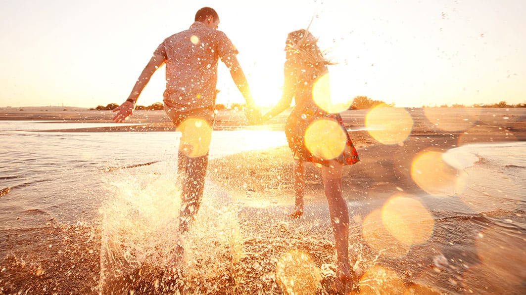 Shown from behind, couple holding hands splashing water as they run along the beach at sunrise