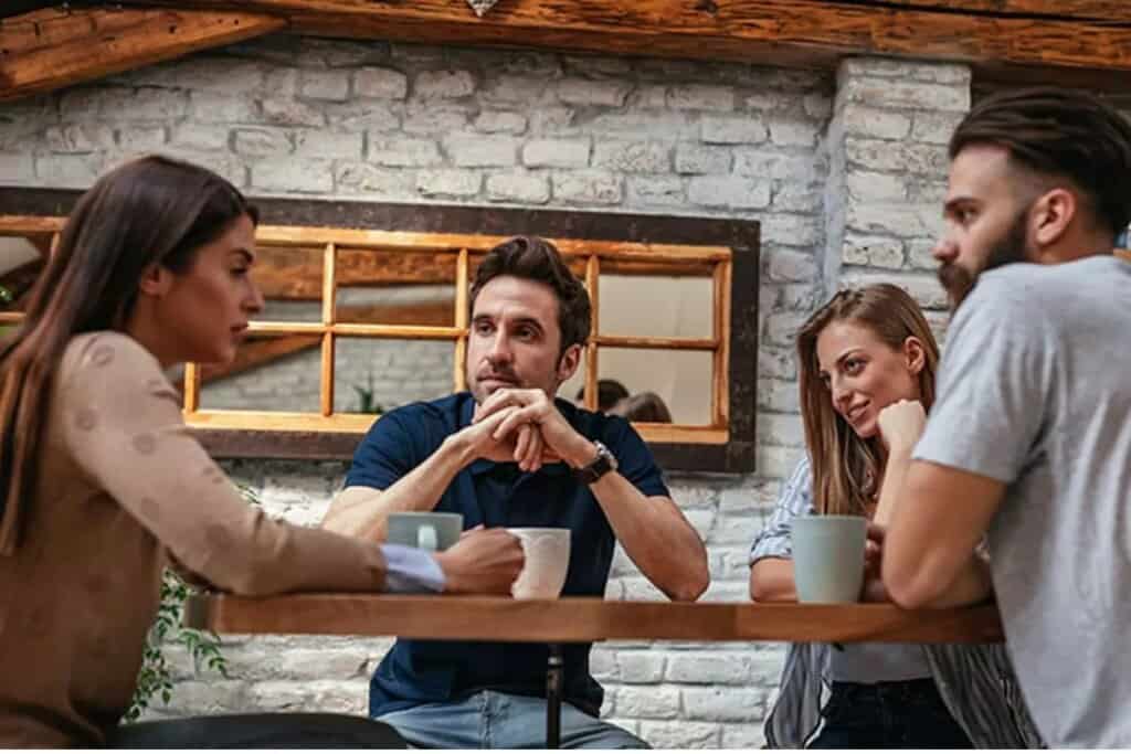 A group of four friends is sitting at a wooden table in a cozy café with exposed brick walls, engaged in a deep conversation. One woman is talking while the others listen attentively, showing different reactions—thoughtfulness, concern, and curiosity. The setting suggests a supportive environment, but it also raises the question of when to talk to a professional counselor instead of just talking to friends.