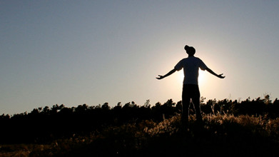 Silhouette of a young person wearing a ball cap and standing on a hill raising his arms in front of a sunrise